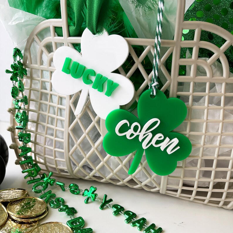 Pu&ograve; includere: Due etichette verdi a forma di trifoglio con le parole "lucky" e "cohen" scritte in bianco. Le etichette sono appese a un cesto di vimini bianco.