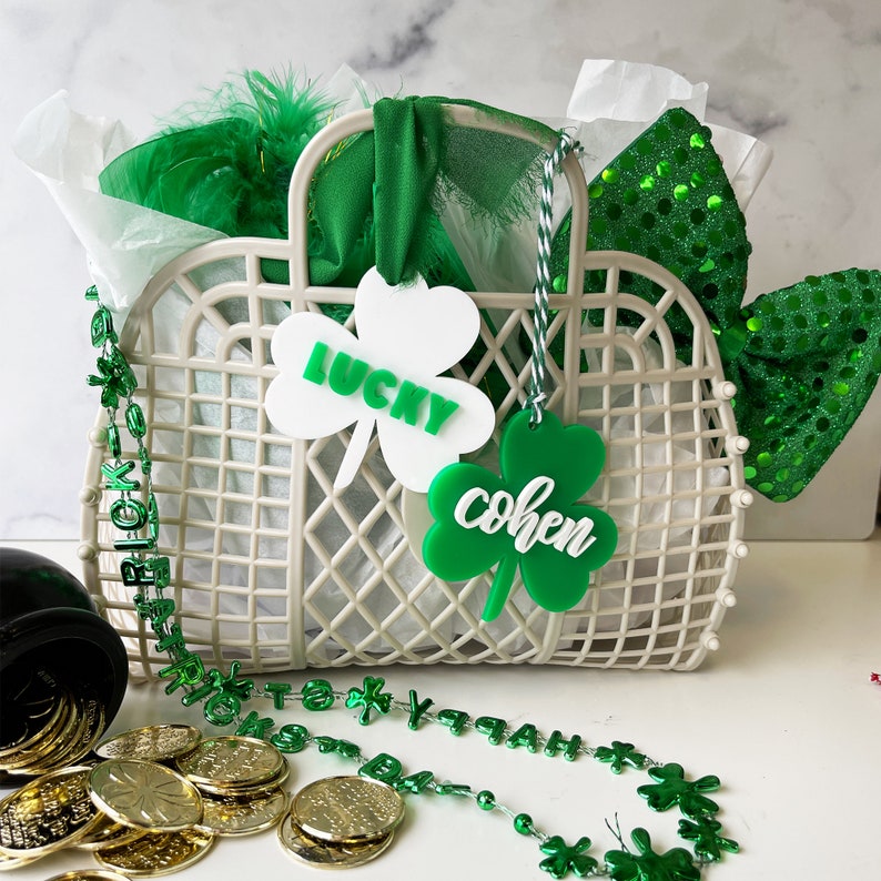 Pu&ograve; includere: Un cesto di vimini bianco pieno di etichette a forma di trifoglio verde e bianco, boa di piume verde e bianco e un fiocco verde scintillante. Le etichette recitano "Lucky" e "Cohen".