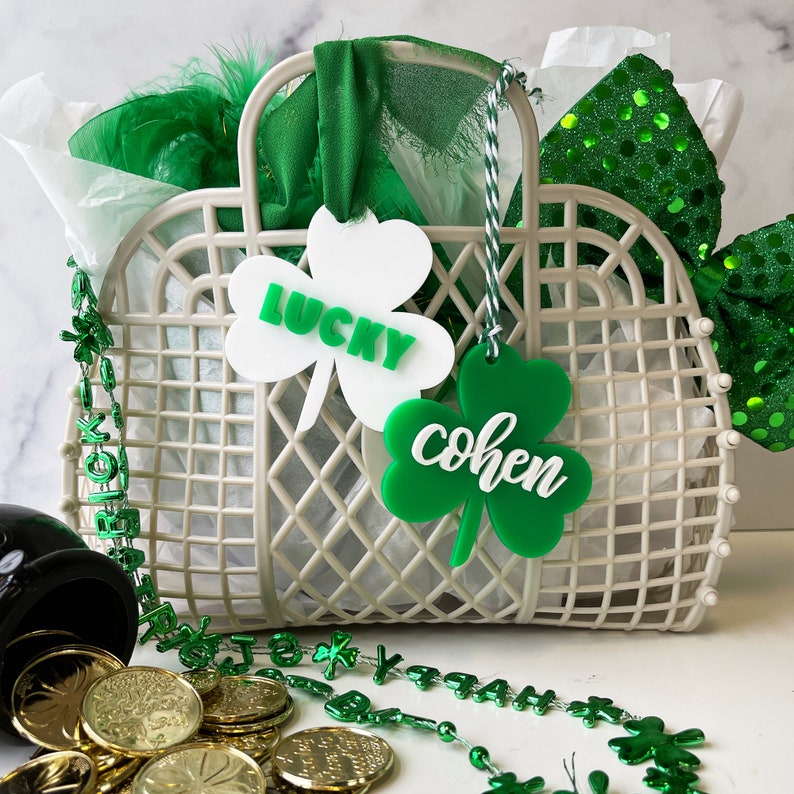 Pu&ograve; includere: Un cesto di vimini bianco con etichette verdi a forma di trifoglio che recitano "Lucky" e "Cohen". Il cesto &egrave; pieno di carta velina verde e decorazioni verdi e bianche. Ci sono monete d'oro e una collana di perline verdi con ciondoli a forma di trifoglio sparsi intorno al cesto.