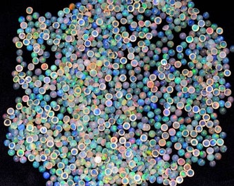 Op de afbeelding: Een verzameling kleine, ronde opalen met een gepolijst, iriserend oppervlak. De opalen vertonen een scala aan kleuren, waaronder blauw, groen, geel en rood, waardoor een glinsterend effect ontstaat. De edelstenen zijn verspreid over een donkere ondergrond.