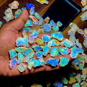 Peut inclure: Une collection de pierres d'opale brutes, affichant un spectre de couleurs comprenant des bleus, des verts et des éclairs irisés. Les opales sont tenues dans une main, avec un plateau noir à proximité. Les pierres précieuses varient en taille et en forme.