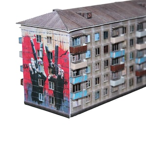 May include: A miniature model of a multi-story apartment building. The building's facade features a red mural of a hand making a peace sign. The building has multiple windows and balconies, with a weathered, aged appearance.
