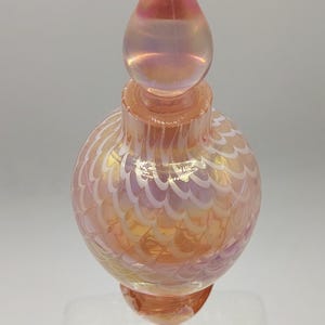 May include: A glass perfume bottle with a spherical body and a tall, pointed stopper. The glass features an iridescent peach and gold finish with a white, swirling feather-like pattern throughout the rounded base and the matching stopper.