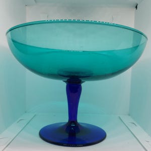 May include: A teal-colored glass serving bowl with a dark blue stem and base. The bowl has a wide, shallow design with a slightly flared rim. The stem is elegantly shaped, and the base is round. This decorative piece is suitable for serving fruit or desserts.