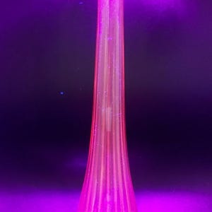 May include: A tall, pink glass vase with a fluted design, illuminated by a purple light. The vase has a flared base and a slender neck. The light source is positioned above the vase, creating a vibrant glow.