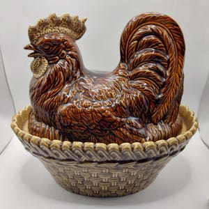 May include: A brown glazed ceramic rooster-shaped lid sitting atop a matching brown basket-weave base. The rooster features detailed feather textures, a prominent comb, and a wattle. The base has a scalloped rim and a woven pattern design.