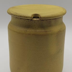 May include: A cylindrical mustard yellow ceramic jar with a matching lid. The jar features a matte, speckled texture throughout its surface. The lid includes a small, semi-circular notch along the rim, designed to accommodate a spoon or utensil.