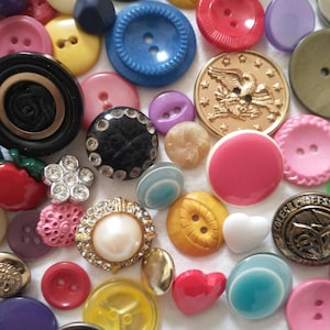 May include: A collection of assorted sewing buttons in various shapes, sizes, and colors. The assortment includes round, heart-shaped, and flower-shaped buttons made of plastic, metal, and faux pearl, featuring textures, embossed eagle designs, and rhinestones.