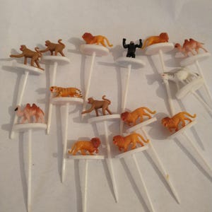 May include: A collection of miniature animal-themed food picks. The picks feature small plastic figurines of lions, monkeys, tigers, camels, and a gorilla. The figurines are attached to white plastic sticks, perfect for appetizers or party snacks.