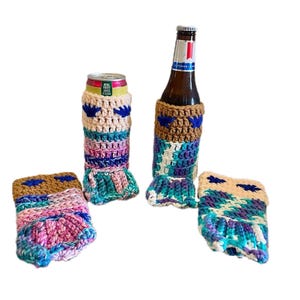 May include: Crocheted drink cozies in shades of brown, blue, pink, and teal. The set includes two can cozies, a bottle cozy, and a coaster. The cozies have a textured design with a brown top and blue accents.