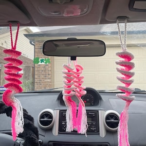 May include: Three handmade car charms hanging from the ceiling of a car. Each charm features a spiral design with alternating pink and white crochet sections, and long white and pink tassels. The charms are suspended in front of the car's dashboard and rearview mirror.