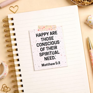 Puede incluir: Un cuaderno de espiral con páginas rayadas, una nota adhesiva con el texto "HAPPY ARE THOSE CONSCIOUS OF THEIR SPIRITUAL NEED. Matthew 5:3" y un bolígrafo con estampado floral. También se ven clips dorados y una cadena decorativa dorada.