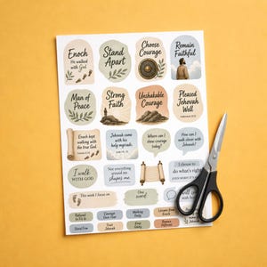 May include: A white sheet of paper featuring various stickers with religious text and illustrations, including phrases like Enoch He walked with God, Stand Apart, and Choose Courage, accompanied by a pair of black-handled metal scissors.