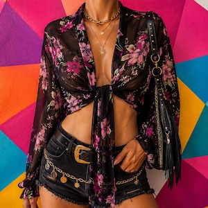 May include: A black sheer long-sleeve blouse with a pink and purple floral pattern, featuring a front tie closure. The blouse is paired with black denim shorts, a black belt with a gold buckle, a gold chain waist accessory, and a black fringe shoulder bag.