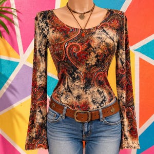 May include: A long-sleeved top featuring a multi-colored paisley pattern in shades of red, black, and tan, paired with blue denim jeans and a brown leather belt with a metal buckle. A layered necklace with a circular pendant is worn.