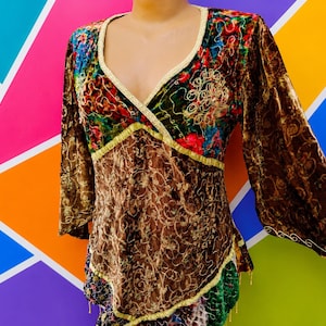 Peut inclure: Un haut à manches longues avec une encolure en V bordée de doré. Le vêtement présente un mélange de velours marron à motifs de volutes dorées et des empiècements en velours floral multicolore. L'ourlet est asymétrique avec des détails dorés.