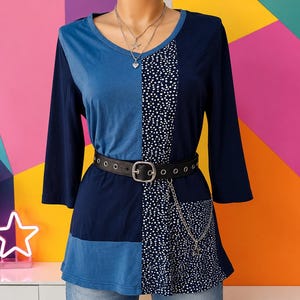 Op de afbeelding: Een top met driekwartmouwen in een color-block ontwerp van marineblauw en lichtblauw, met een verticaal paneel met witte stippen op een donkere achtergrond. Het kledingstuk is getailleerd met een zwarte riem met oogjes en een zilveren ketting.