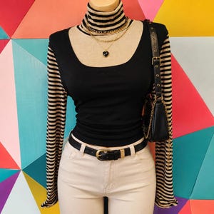 Puede incluir: Una blusa de manga larga con cuerpo negro de cuello cuadrado sobre un cuello alto de rayas beige y negras, combinada con pantalones claros, cinturón negro con hebilla dorada, bolso de hombro negro y collar dorado en capas con un colgante de corazón.