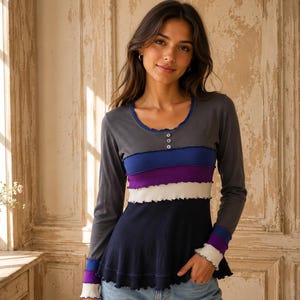 Può includere: Una maglia a maniche lunghe con scollo rotondo, tre bottoni e un design a blocchi di colore. Il capo presenta fasce orizzontali nei toni del grigio antracite, blu, viola, bianco e blu navy con bordi arricciati sui polsini e sull'orlo.