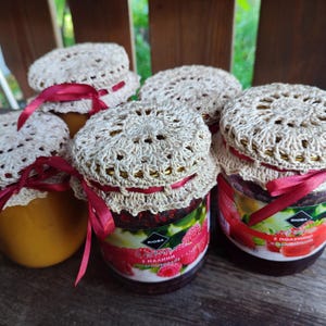 Op de afbeelding: Verschillende glazen potten met jam, elk afgedekt met een crèmekleurig gehaakt kleedje en vastgebonden met een rood lint. De potten bevatten een gele jam en een rode jam met een frambozenlabel.