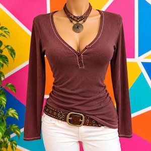 Op de afbeelding: Een bordeauxrood henley-shirt met lange mouwen, een V-hals en drie kleine knoopjes. Het kledingstuk heeft zichtbare lichtgekleurde stiksels langs de halslijn en de zoom. Het wordt gecombineerd met een bruine leren riem met metalen studs.
