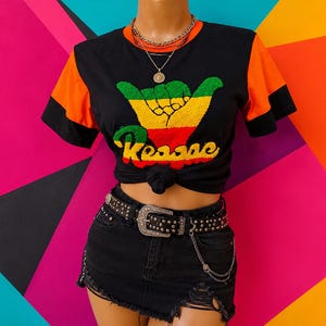 May include: A black t-shirt with orange sleeves and a graphic print of a hand making a shaka sign in green, yellow, and red colours with the text Reggae. The shirt is paired with a black denim skirt featuring distressed details and a studded belt.