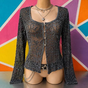 May include: A dark grey long-sleeve button-down cardigan featuring an intricate floral lace pattern with a sheer mesh texture. The garment has a scoop neckline and is displayed on a mannequin wearing a layered silver necklace and a black star belt.