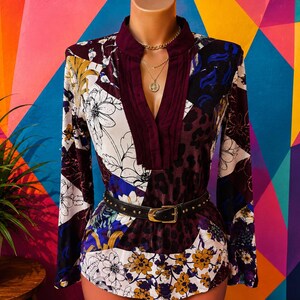 Pode incluir: Uma blusa de manga comprida com design de patchwork, apresentando padrões florais em branco, amarelo e azul, juntamente com secções de estampado de leopardo em roxo escuro. A peça tem um decote profundo em bordô e um cinto preto com tachas.