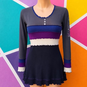 May include: A long-sleeve top featuring a color-blocked design with horizontal stripes in dark gray, blue, purple, and cream. The garment has a scoop neckline with three small buttons, long sleeves with matching striped cuffs, and a ruffled hem.