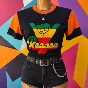 May include: A black t-shirt with orange sleeves and a black trim, featuring a textured, fuzzy graphic of a hand making a shaka sign above the word Reggae in red, yellow, and green. The shirt is paired with black denim shorts and a black studded belt.