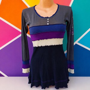 Puede incluir: Una camiseta de manga larga con cuello redondo y tres botones pequeños. El cuerpo está formado por paneles horizontales en tonos gris, azul, morado y blanco con costuras sin rematar. Las mangas y la parte inferior son de color azul marino.