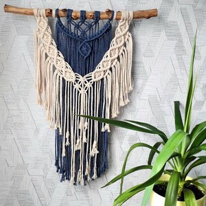 May include: A blue and white macrame wall hanging with a geometric pattern. The hanging is made of cotton cord and has a fringe at the bottom.