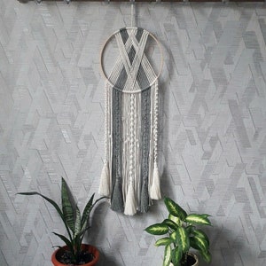 Könnte beinhalten: Ein grauer und weißer Makramee-Traumfänger mit einem Holzreifen und Quasten, die von unten hängen. Der Traumfänger hängt an einer weißen Wand mit einem geometrischen Muster.