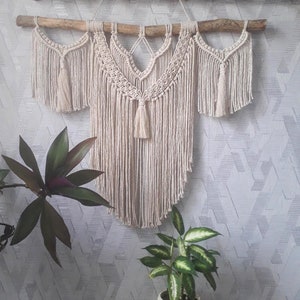 Pode incluir: Uma tapeçaria de macramé de parede com um ramo de madeira natural no topo. A tapeçaria é feita de fio de cor creme e tem uma franja na parte inferior. A tapeçaria tem a forma de uma lágrima.