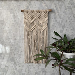 May include: A beige macrame wall hanging with a geometric pattern. The hanging is made of cotton cord and has a wooden dowel at the top.