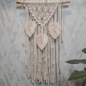 Peut inclure: Une suspension murale en macram&eacute; beige avec un motif g&eacute;om&eacute;trique et trois formes de feuilles. La suspension est faite de cordon de coton naturel et est fix&eacute;e &agrave; une baguette en bois.