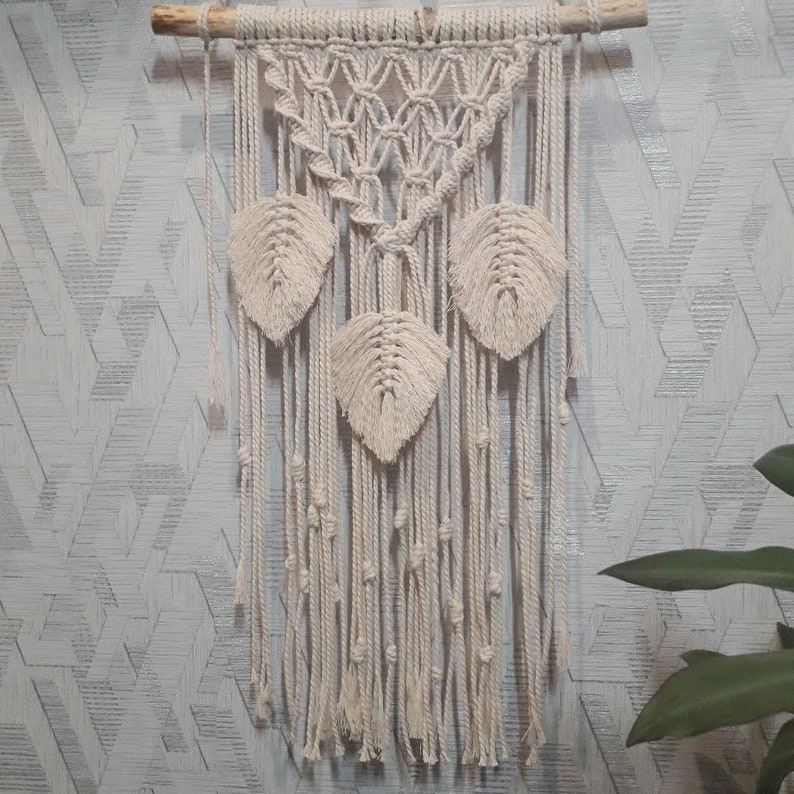Peut inclure: Une suspension murale en macram&eacute; beige avec un motif g&eacute;om&eacute;trique et trois formes de feuilles. La suspension est faite de cordon de coton naturel et est fix&eacute;e &agrave; une baguette en bois.