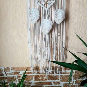 Peut inclure: Une suspension murale en macram&eacute; blanche avec un motif g&eacute;om&eacute;trique et trois pompons blancs en forme de plumes. La suspension est fix&eacute;e &agrave; une branche de bois naturel.