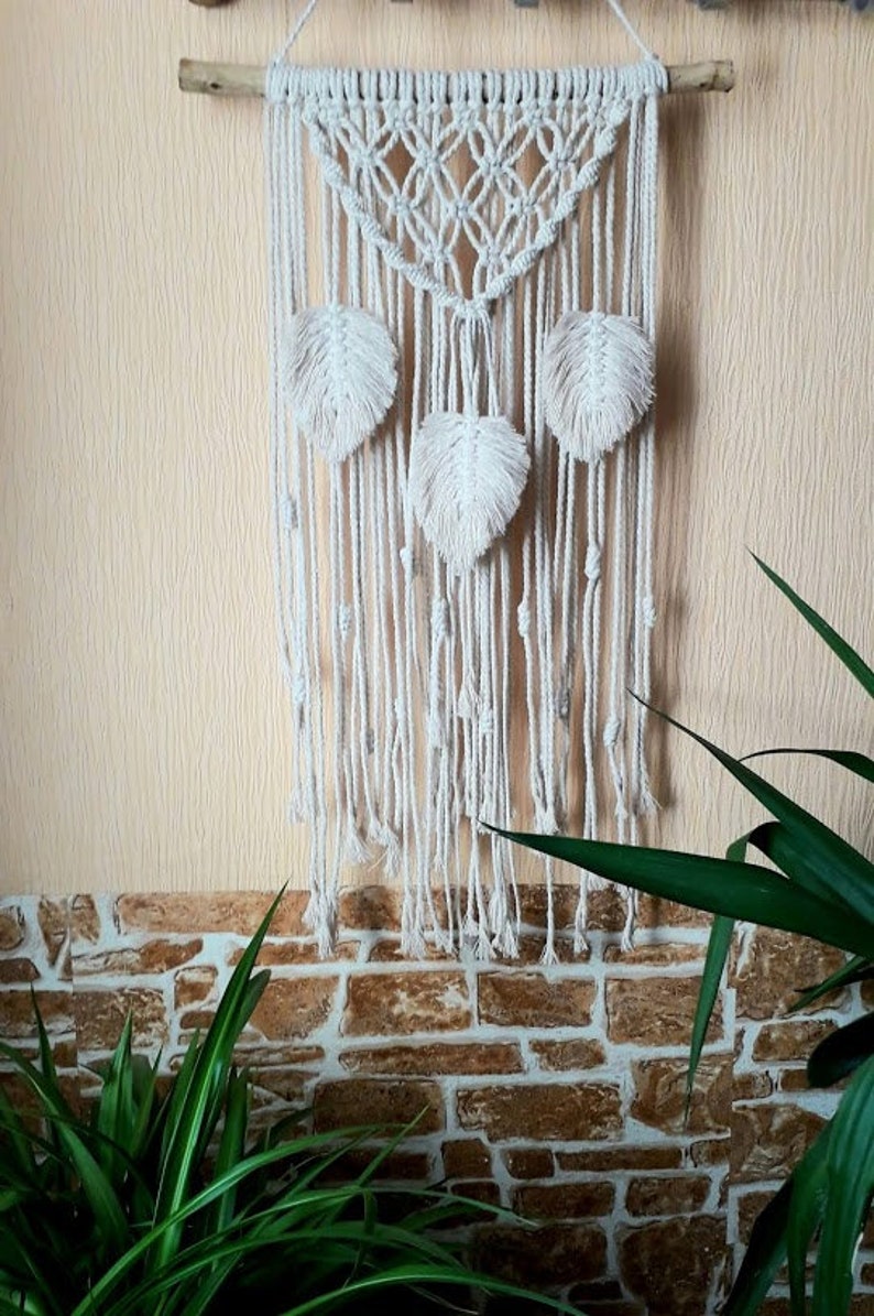 Peut inclure: Une suspension murale en macram&eacute; blanche avec un motif g&eacute;om&eacute;trique et trois pompons blancs en forme de plumes. La suspension est fix&eacute;e &agrave; une branche de bois naturel.