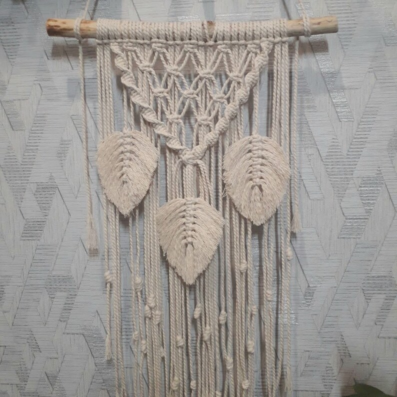 Peut inclure: Une suspension murale en macram&eacute; blanche avec un motif triangulaire et trois formes de feuilles. La suspension est faite de cordon de coton naturel et a un b&acirc;tonnet en bois en haut.