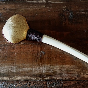 May include: A handcrafted club featuring a light tan, rounded head and a long, off-white handle. The head is wrapped with a dark brown material, and the handle is smooth. The club is likely made of wood and leather.