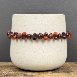 May include: A red and brown beaded bracelet with a knotted cord. The beads are a mix of red tiger's eye and garnet.