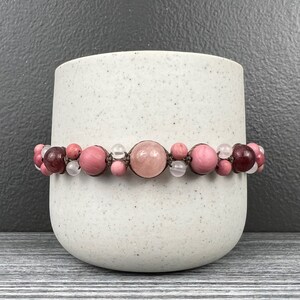 May include: A pink and brown beaded bracelet with a large pink gemstone in the center. The bracelet is on a white ceramic surface.