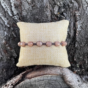 May include: A light pink gemstone bracelet with brown cord and knots. The bracelet is displayed on a beige fabric cushion.