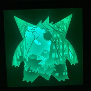 May include: A green glow-in-the-dark papercraft of a spooky ghost with trees and a building in the background.