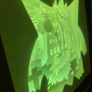 May include: A green glow-in-the-dark papercraft scene depicting a spooky Halloween graveyard with a haunted house, a cross, and a tree with a spider web.