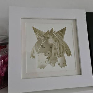 May include: A white framed picture with a white paper cutout of a cartoon character inside. The character is a white wolf with a black nose and eyes. The wolf is surrounded by a white forest scene.