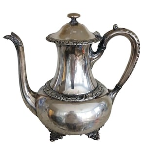 May include: A silver-colored teapot with a curved spout and handle. The teapot has a lid with a small knob and is decorated with ornate detailing around the base and lid. The teapot sits on three small feet.