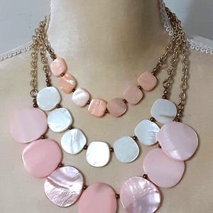May include: A multi-strand necklace featuring three layers of flat, circular, and square-shaped shell beads in shades of pale pink, peach, and iridescent white. The beads are strung on gold-toned metal chains with small metallic spacer beads.