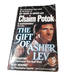 May include: A paperback book titled The Gift of Asher Lev by Chaim Potok. The black cover features a portrait of a man and a child, with white text stating The extraordinary new novel by the author of My Name is Asher Lev and A Masterwork.